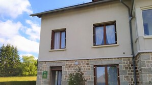 A two-story building with a stone facade, a door labeled 'Gite,' and a small garden area with a tree and flowers. - Gîte Le Toura 2/4 persons (SAINT-ALBAN-SUR-LIMAGNOLE)