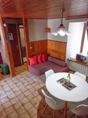 Ein gemütliches Zimmer mit Holzbalkendecke, einer Bank mit roten Kissen, einem weißen Tisch mit Stühlen und einer kleinen Küchenzeile mit einer Mikrowelle.