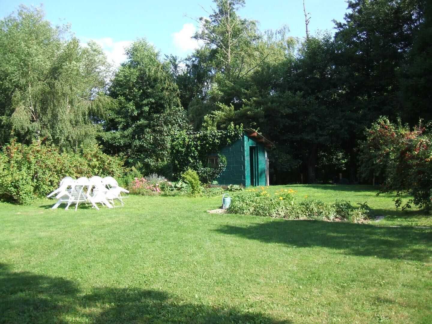 Ein grüner Rasen mit einem kleinen Schuppen, weißen Stühlen und dichter Baumgruppe im Hintergrund.