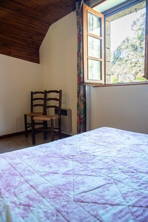 Ein Zimmer mit einem Bett, einem Sessel, einem Fenster mit Blick auf eine felsige Landschaft und einer Holzbalkendecke.