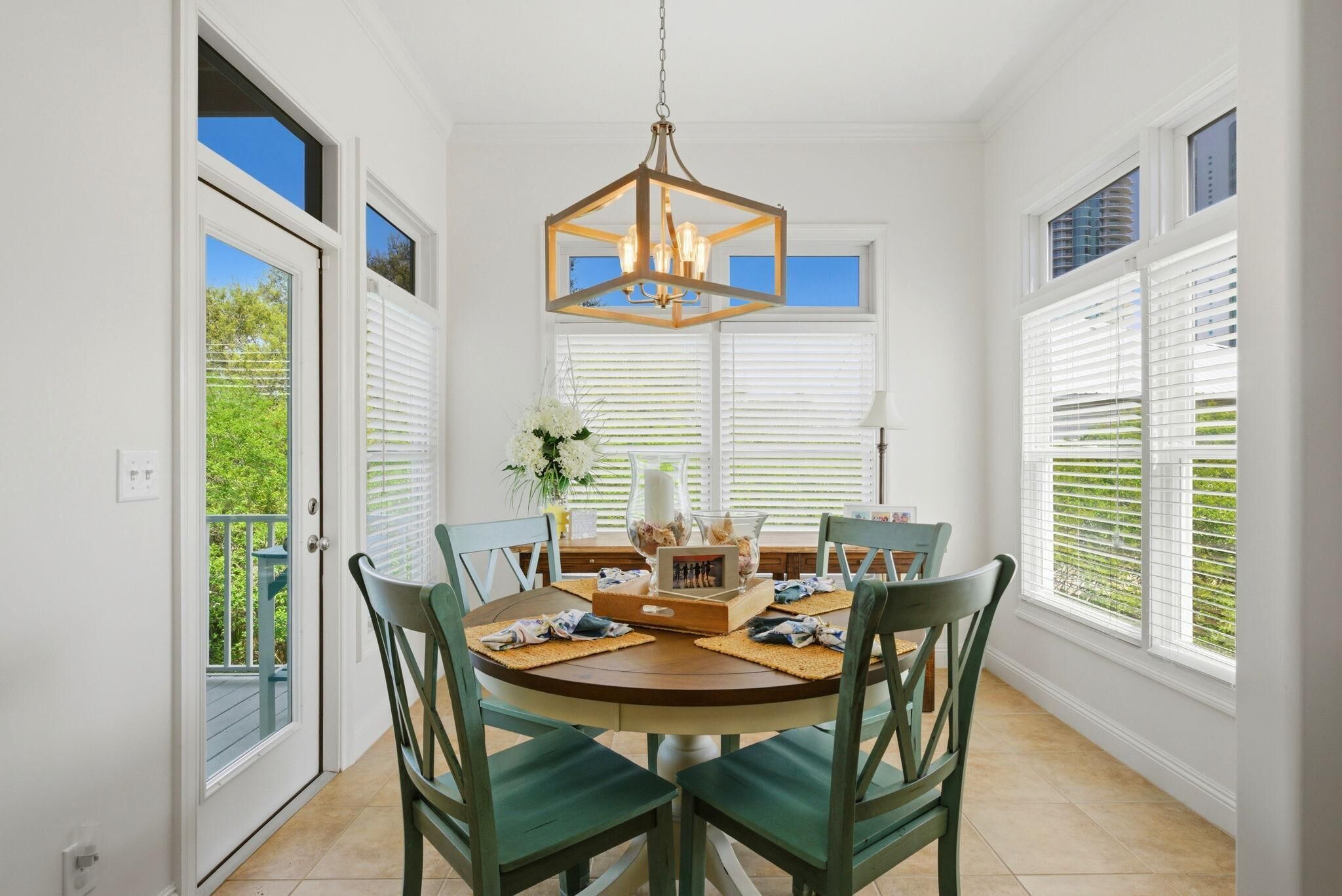 A dining area with a round table, green chairs, a chandelier, and a view through large windows.