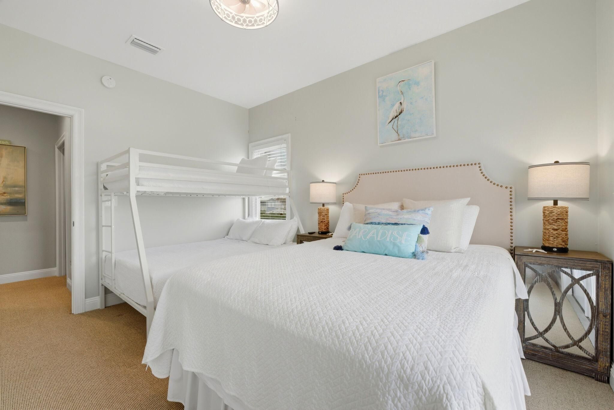 A bedroom with a bunk bed, a large bed with a quilt, two bedside lamps, and a painting on the wall.