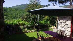 Een terras met een tafel en stoelen, een stenen muur en uitzicht op een weelderige tuin en heuvels.