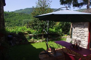 A patio with a table and chairs, a stone wall, and a view of a lush garden and hills. - Chez MamieThé - ISPAGNAC (ISPAGNAC)