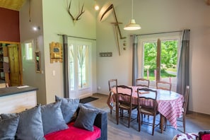 A room with a dining table set for four, a sofa with cushions, a large window with curtains, and a wall-mounted deer skull. - La Source du Cellier 4/6 persons (SAINT-JEAN-LA-FOUILLOUSE)