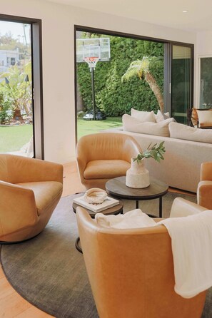 A modern living room with leather sofas, a coffee table, and a view of a basketball hoop and greenery outside.