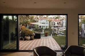 A modern interior with large sliding glass doors leading to an outdoor patio area with a patterned floor, potted plants, and a view of neighboring houses.