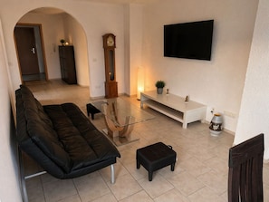 A living room with a sofa, ottoman, glass coffee table, TV, and a clock. - Vacation apartment / apartment for fitters / parking lot / WLAN / kitchen / 120QM (Urbar)