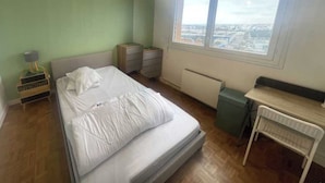 A bedroom with a bed, bedside tables, a chair, a radiator, and a window with a city view. - Private Room in a Shared Flat - Villeurbanne (Villeurbanne)