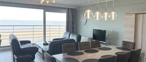 A modern living room with a dining area, large windows, and a view of the beach.