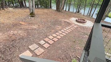 Une terrasse avec un chemin de pierre menant à un foyer extérieur, entourée d’une zone boisée et d’un plan d’eau.