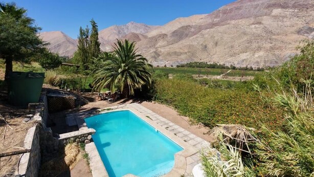 Una piscina con montañas al fondo, rodeada de vegetación y un muro de piedra.