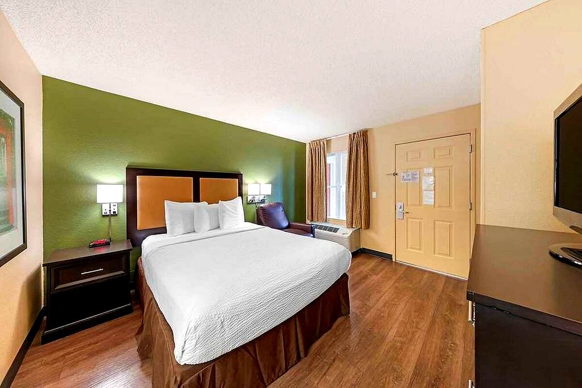 A hotel room with a bed, a nightstand, a TV, a door, and a window with curtains.