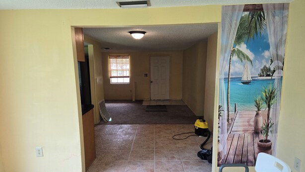 A room with a tiled floor, a door, a window with blinds, a ceiling vent, a yellow vacuum cleaner, and a chair. - 2-bedroom house in peaceful Port Richey with cool AC (Port Richey)