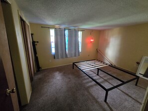 Ein Zimmer mit Bettgestell, einem Vorhangfenster, einer Tür und einer Wandleuchte.