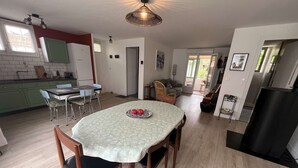 Een eethoek met een tafel, stoelen en een bord eten. Er is een keuken met groene kastjes en een raam.