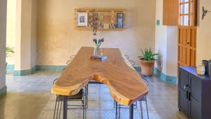 Un'area pranzo con un tavolo di legno lungo, due sedie, un pavimento piastrellato, un soffitto con travi rosse e una mensola a parete con quadri e una pianta.