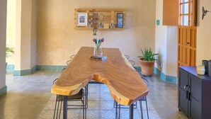 Een eetgedeelte met een lange houten tafel, twee stoelen, een betegelde vloer, een plafond met rode balken en een wandplank met foto's en een plant.