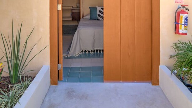 A wooden door with a barred window, a bed with a blue pillow, and a fire extinguisher on the wall. - OX | Private Studio | Casa Na`ay | Boutique Hotel (Mérida)