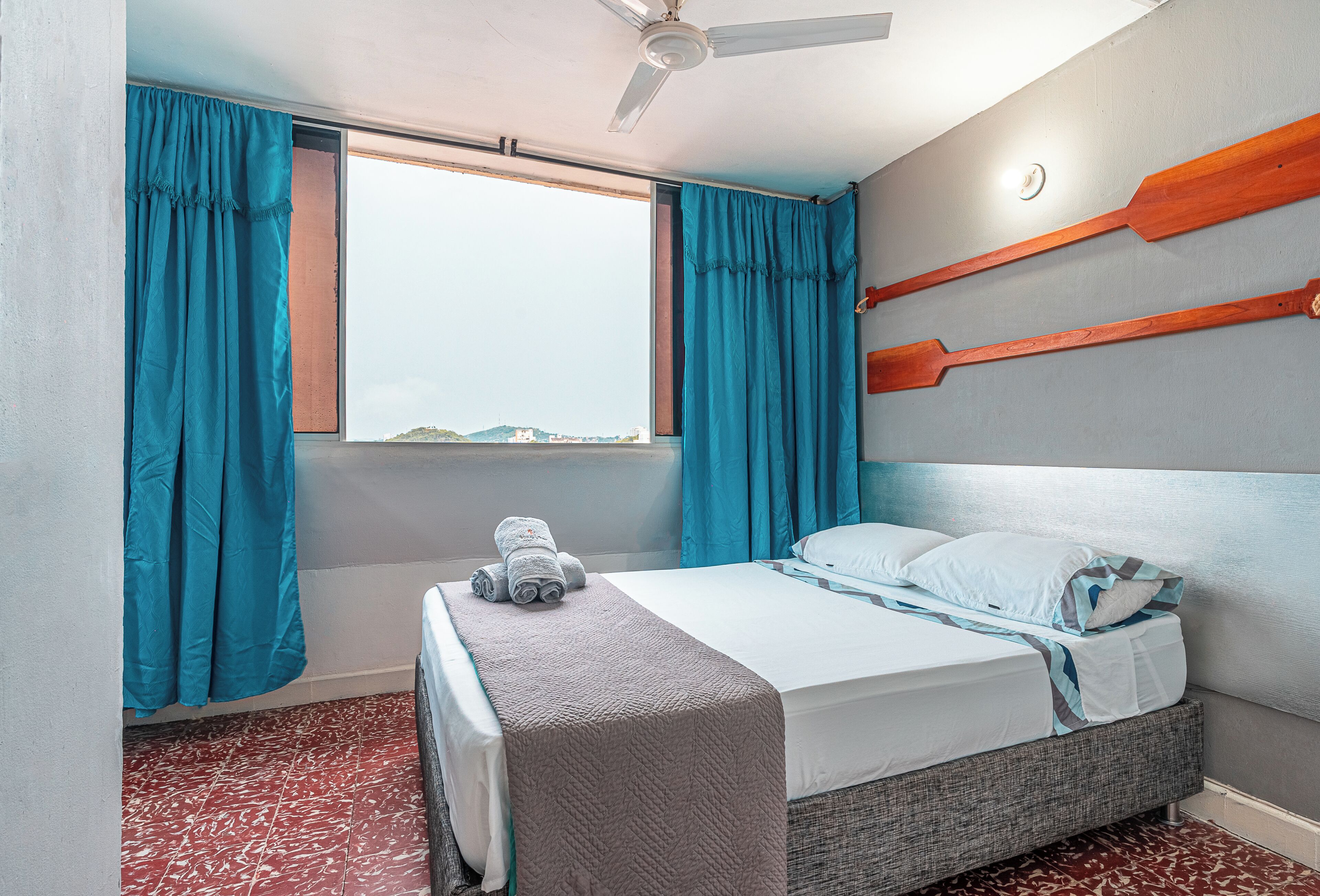 A hotel room with a bed, a window with curtains, a ceiling fan, and a view of the outside.