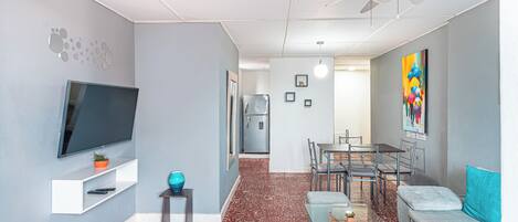 Una sala de estar moderna con una televisión de pantalla plana, un ventilador de techo, una zona de comedor con mesa y sillas, y una cocina al fondo.