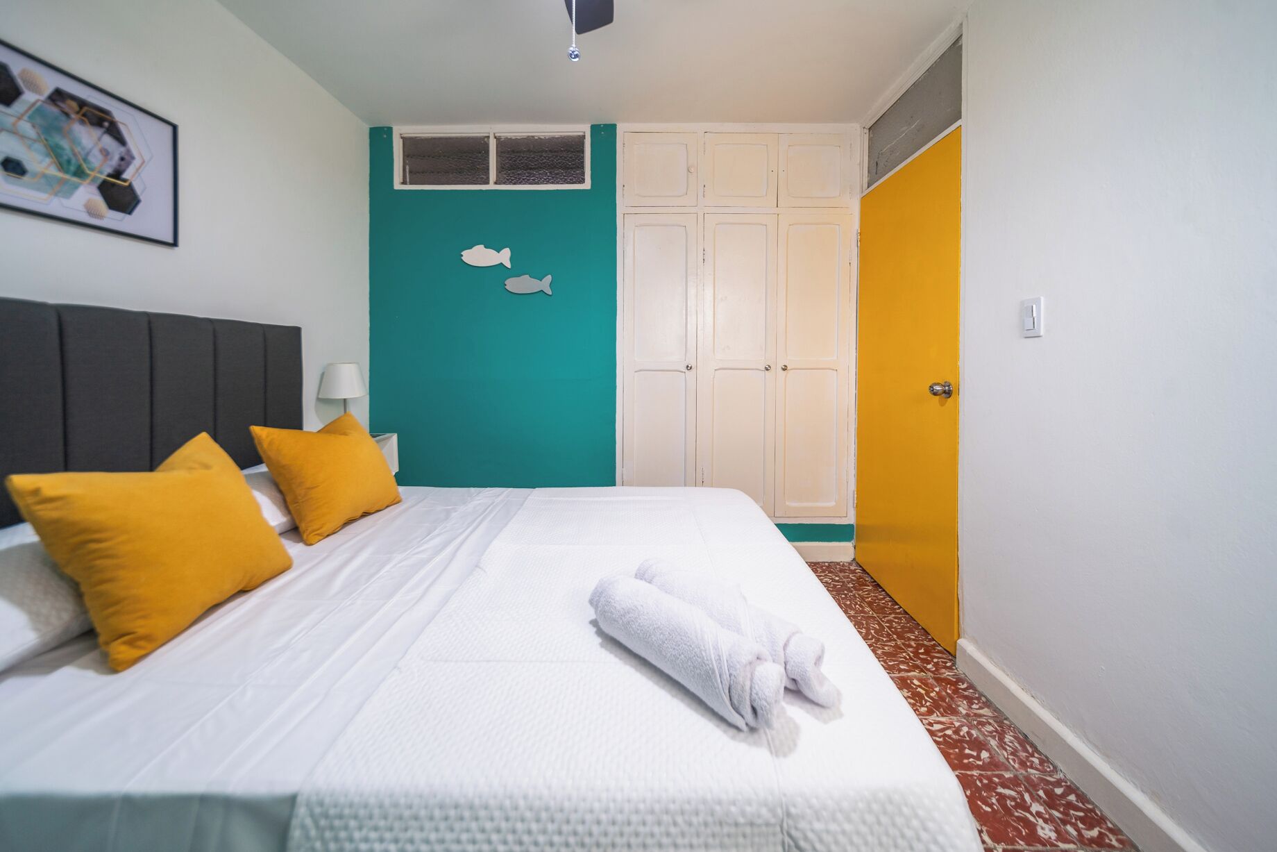 A hotel room with a bed, two yellow pillows, a white towel, a teal accent wall, a white wardrobe, and a yellow door.