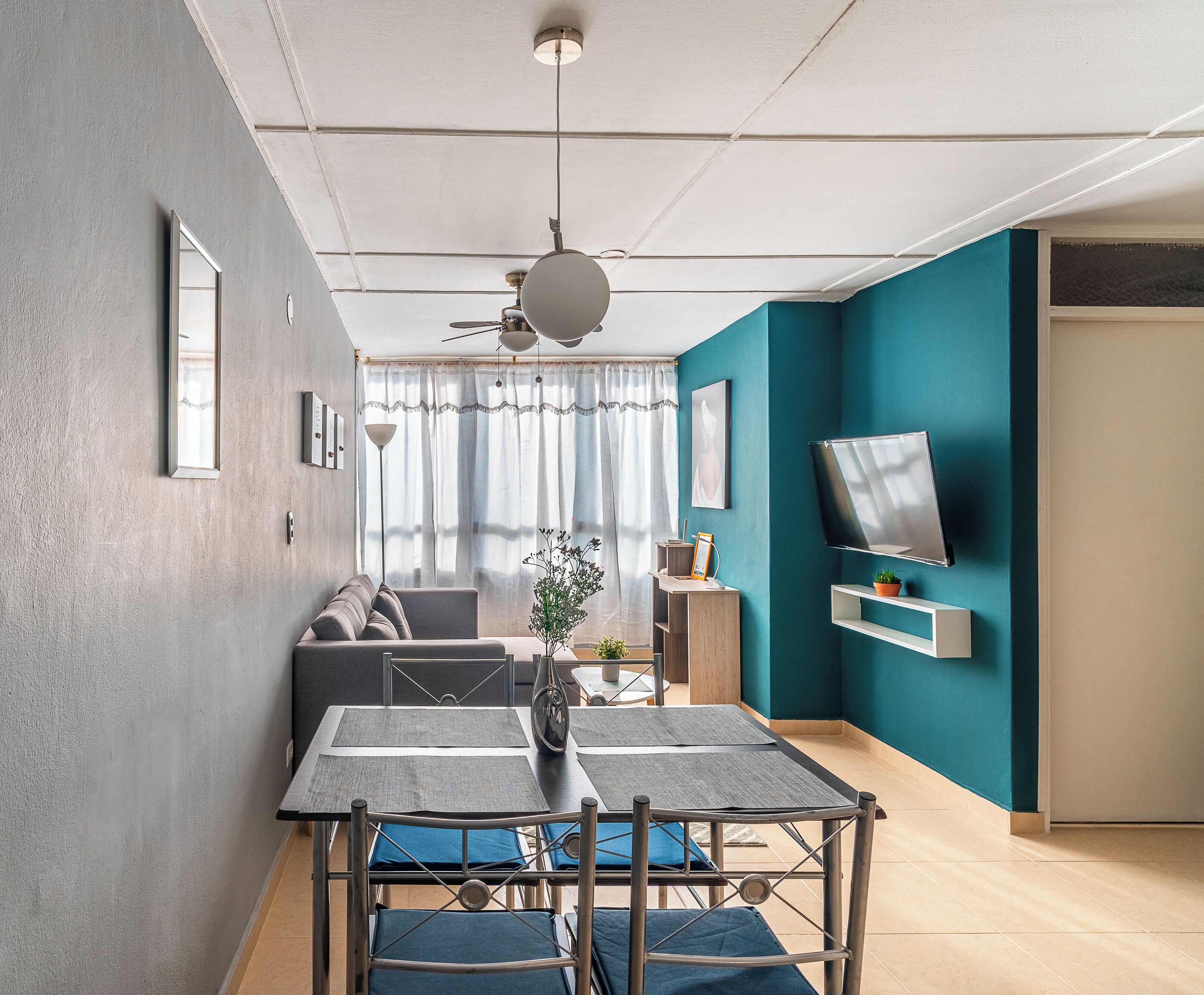 A modern living room with a sofa, a dining table with chairs, a TV mounted on the wall, and a ceiling fan.