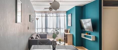 A modern living room with a sofa, a dining table with chairs, a TV mounted on the wall, and a ceiling fan.