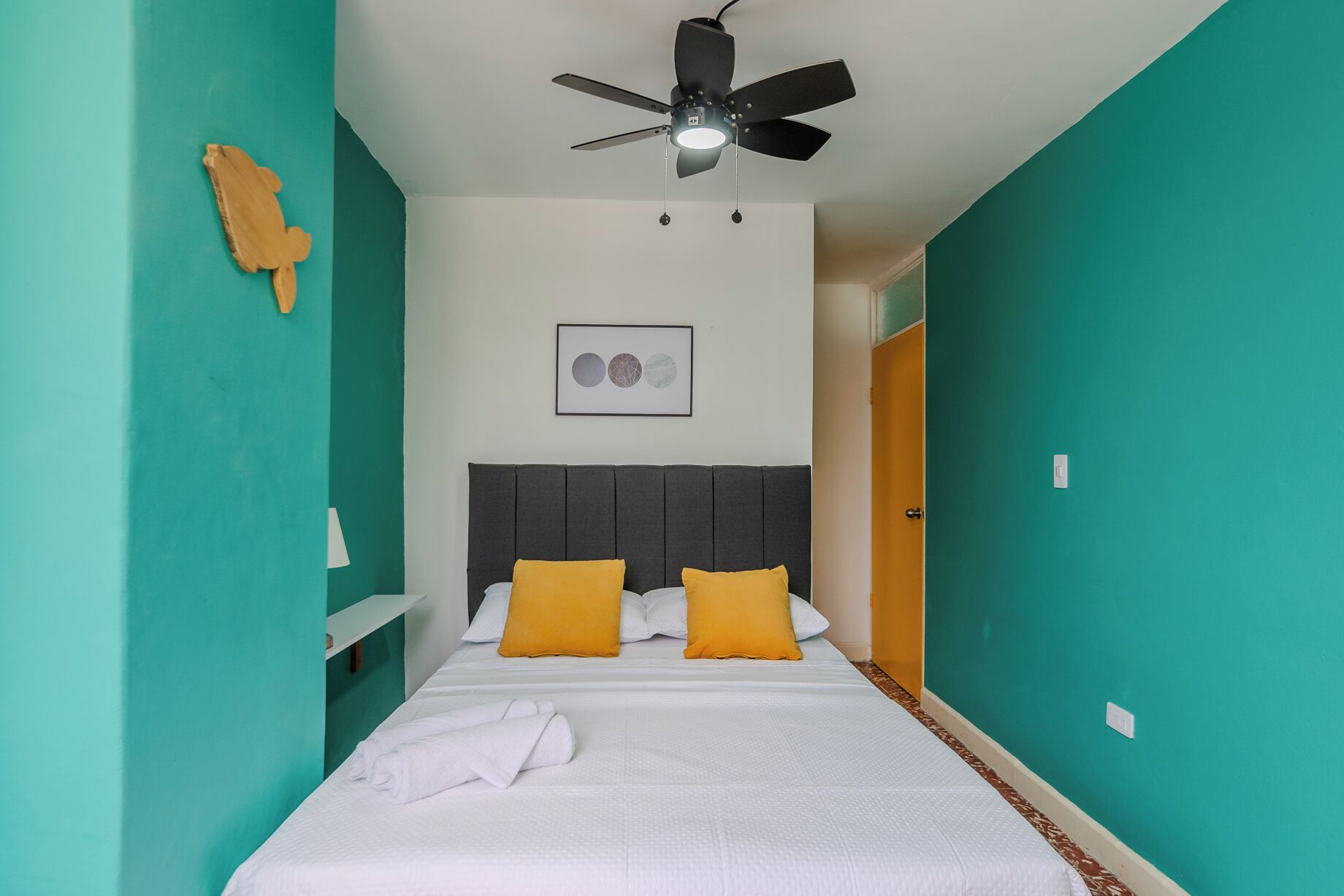 A bedroom with teal walls, a bed with white bedding and two yellow pillows, a ceiling fan, and a framed picture on the wall.
