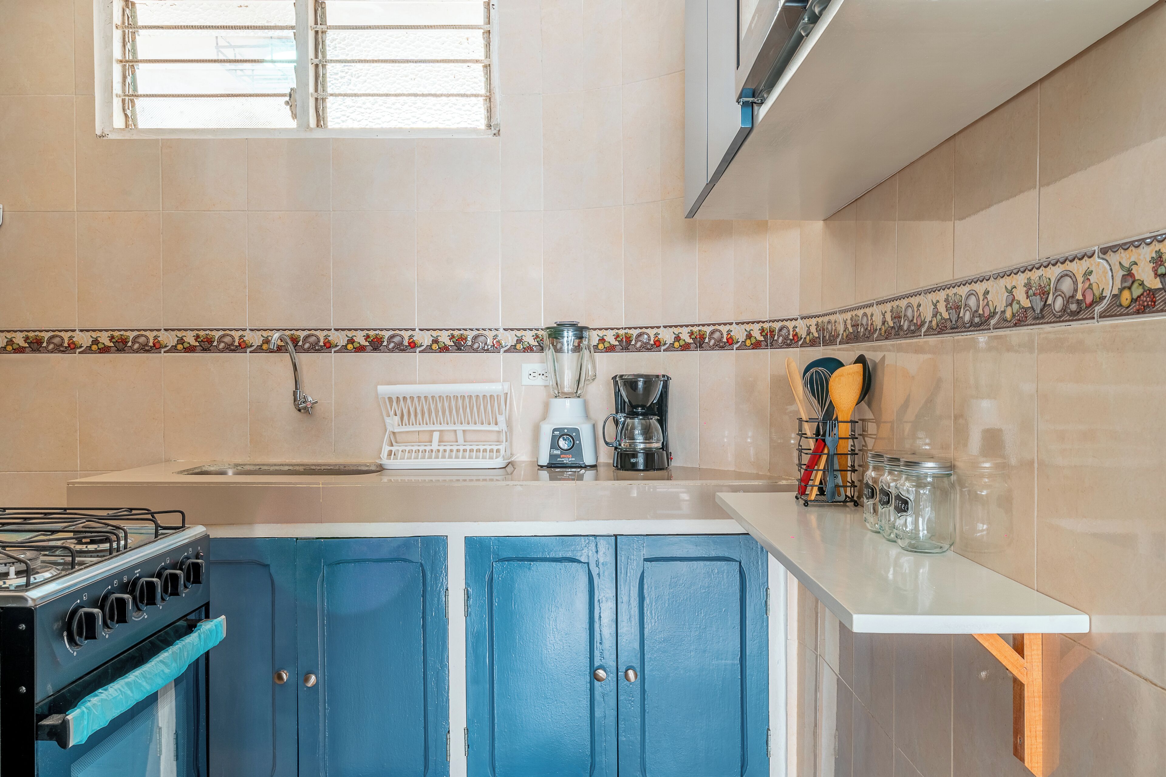 A kitchen with blue cabinets, a stove, a sink, a window, and a countertop with kitchen utensils and jars.