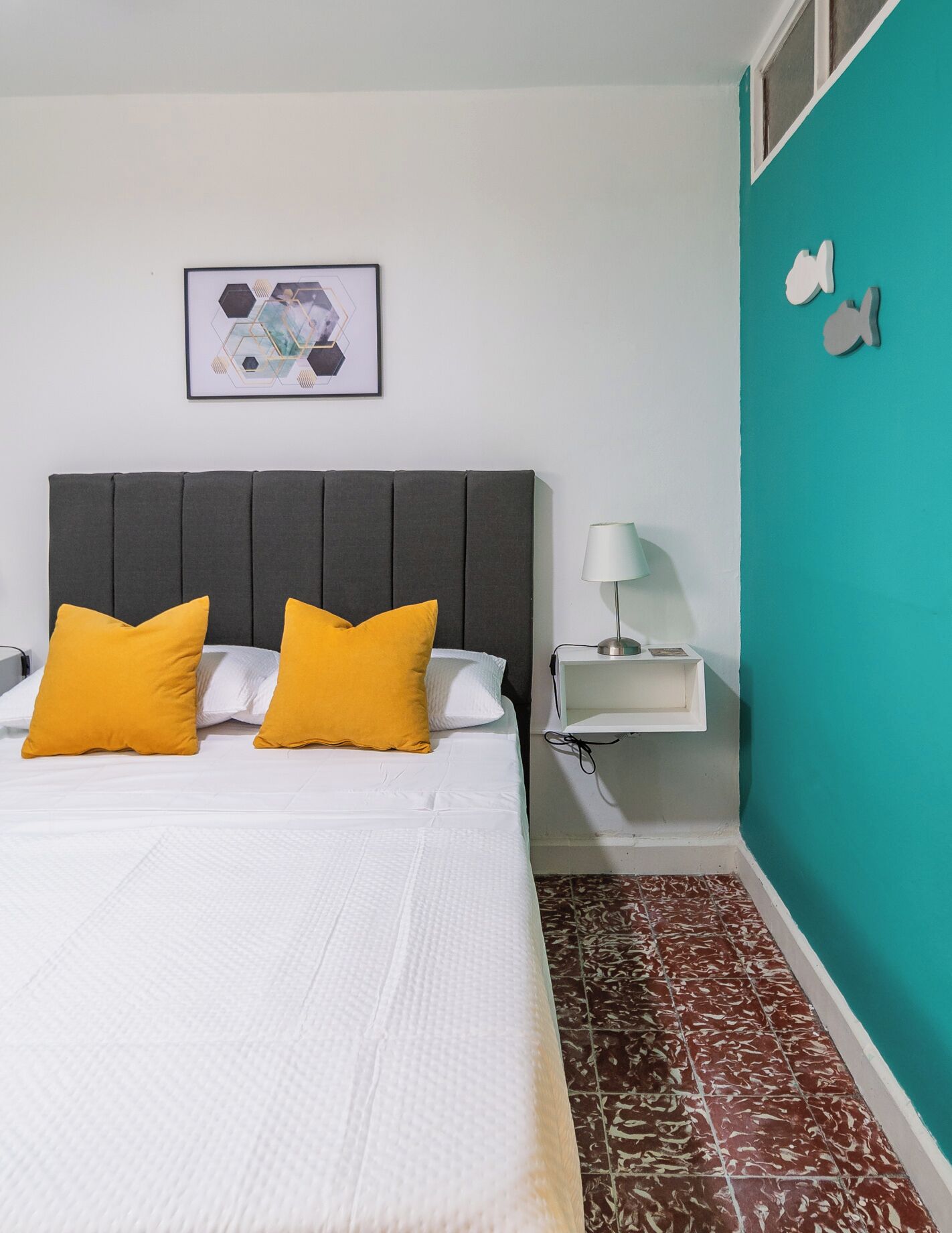 A bed with two yellow pillows, a grey headboard, a white bedside table with a lamp, and a wall-mounted picture.