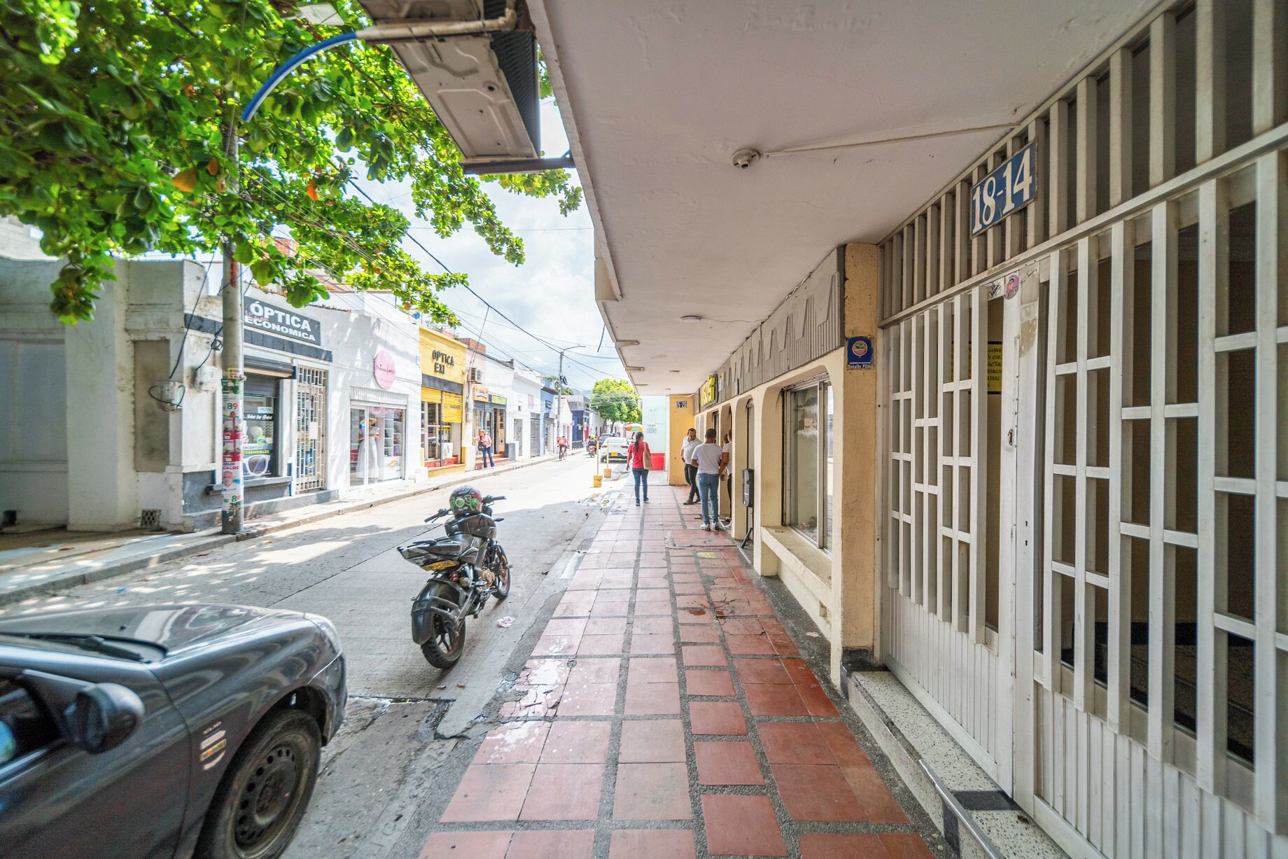 A street with a motorcycle parked on the sidewalk, a building with barred windows, and a sign with the number 18-14.
