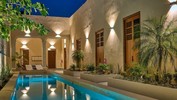Une piscine à l’eau d’un bleu limpide, entourée d’un bâtiment doté de nombreuses portes et fenêtres, et dont l’extérieur est bien éclairé.