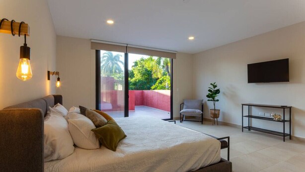 A bedroom with a bed, a chair, a TV, and a balcony with a view of greenery.