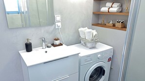 A modern bathroom with a white sink, a washing machine, a mirror, and a shelf with towels and toiletries. - Blue Marsis Apt Paros Escape (Paros)