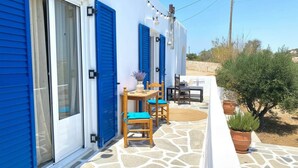 A building with blue shutters, a stone-paved patio, and a small table with chairs. - Blue Marsis Apt Paros Escape (Paros)