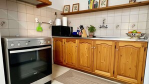 A kitchen with a stove, oven, wooden cabinets, a sink, and a shelf with books and a bowl of fruit. - Blue Marsis Apt Paros Escape (Paros)
