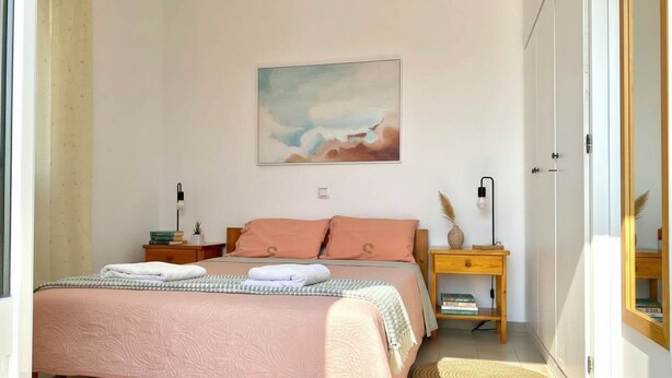 A bedroom with a bed, two bedside tables, a nightstand, a wall-mounted lamp, and a painting. - Blue Marsis Apt Paros Escape (Paros)