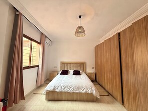 Ein Schlafzimmer mit einem Bett, einer Hängelampe, einem Fenster mit Vorhängen und einem Holzschrank.