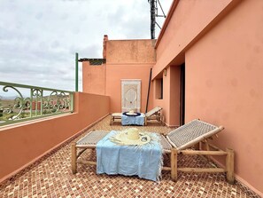 Ein Balkon mit Fliesenboden, zwei Korbstühle, einem kleinen Tisch mit blauer Tischdecke und einem Wandbehang.