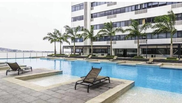 Ein Swimmingpool mit Liegestühlen, Palmen und einem modernen Mehrstöckengebäude.