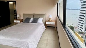 Ein Hotelzimmer mit einem Bett, zwei Nachttischen, einem Fenster mit Blick auf das Wasser und einer Tür.
