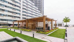 Ein moderner Außenbereich mit Holz-Pergola, umgeben von gepflegtem Rasen und Blumenbeeten, vor einem hohen Wohnhaus.