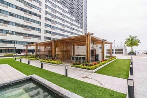 A modern outdoor seating area with a wooden pergola, surrounded by well-maintained grass and flower beds, in front of a tall residential building. - Cozy apartment in the heart of Guayaquil (Guayaquil)