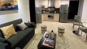 Ein Wohnzimmer mit Sofa, Couchtisch und Hocker, eine Küche mit Schränken und Geräten, sowie ein Bild an der Wand.