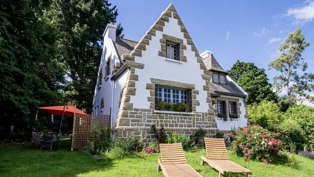 Ein Steinhaus mit einem dreieckigen Dach, ein Garten mit zwei Holzliegen und einem roten Sonnenschirm.