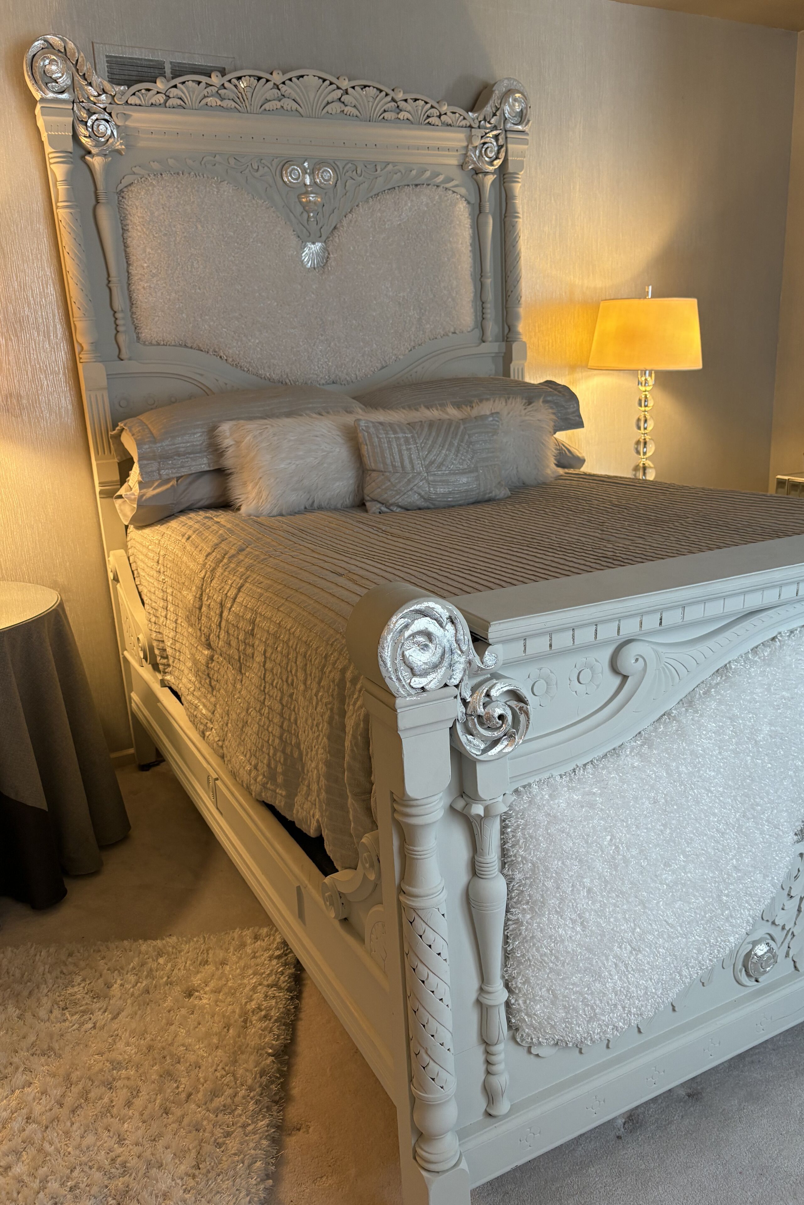 A bed with ornate headboard and footboard, a bedside lamp, and a fluffy rug.