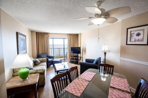 A hotel room with a dining table, chairs, a sofa, a TV, a ceiling fan, and a window with curtains. - Beachfront 3BR Retreat w/ Balcony & Pool (Murrells Inlet)