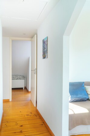 Un couloir étroit au plancher en bois, une porte blanche et, au fond, un lit recouvert d’une literie à motifs.