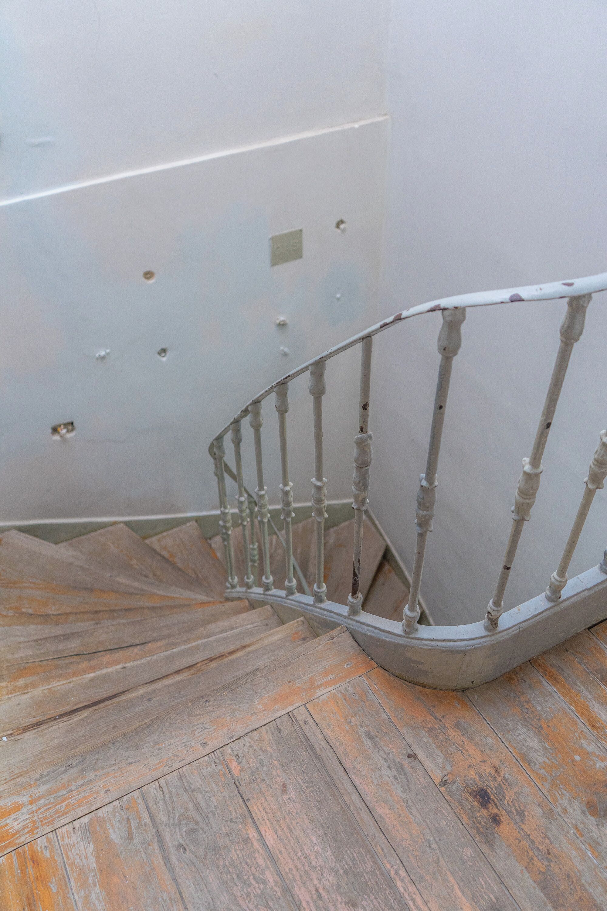 A spiral staircase with a metal railing, wooden steps, and a plain wall with some holes.